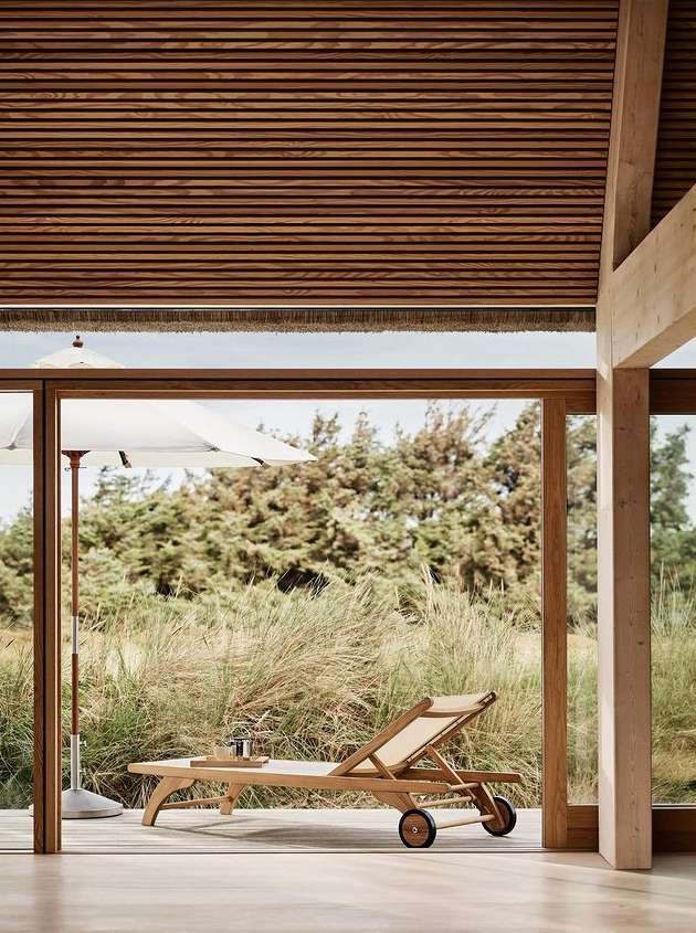 A tan lounge chair sits on a wooden porch in a modern indoor-outdoor space, an umbrella and trees are visible through the window.
