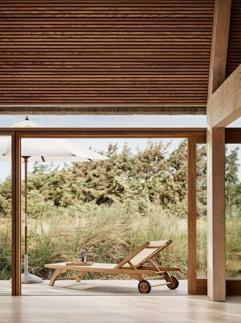 A tan lounge chair sits on a wooden porch in a modern indoor-outdoor space, an umbrella and trees are visible through the window.