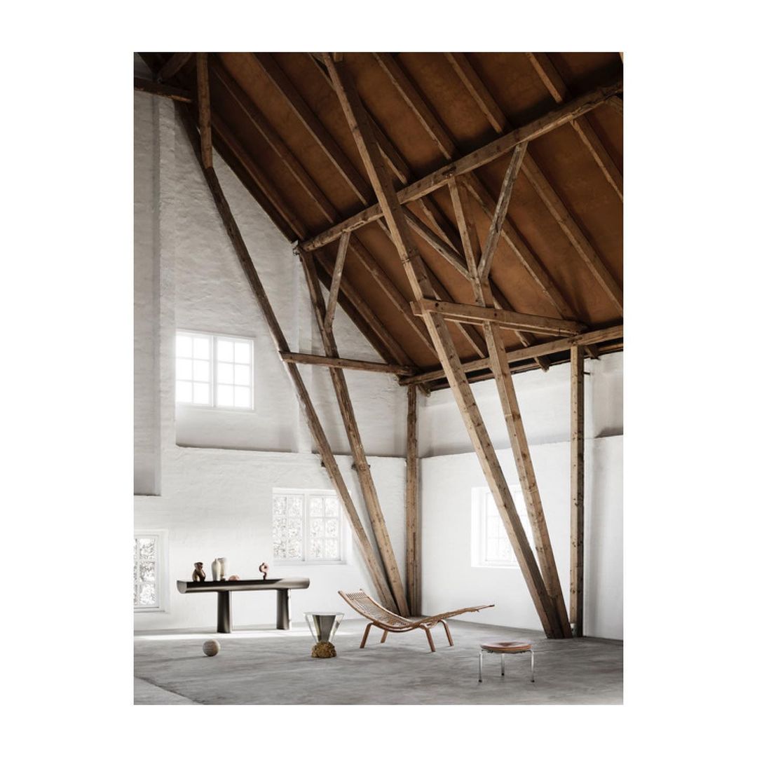 Spacious room showcasing modern furniture, including a wood lounge chair and sculptural table, under high ceilings with exposed beams.