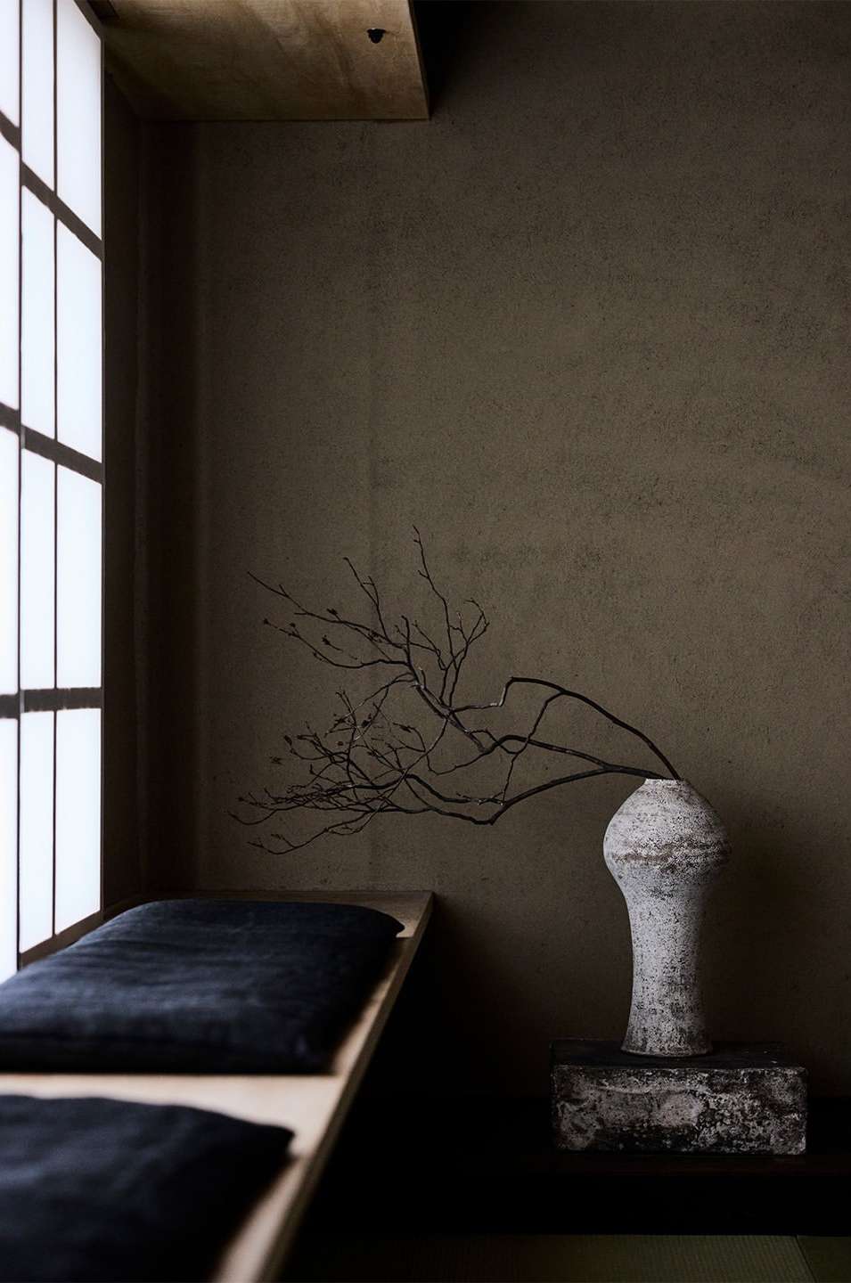 Interior with vase, bare branches, and a cushioned bench near a window, creating a minimalist, neutral-toned, tranquil aesthetic.