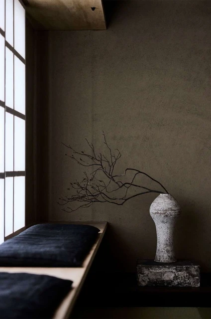 Interior with vase, bare branches, and a cushioned bench near a window, creating a minimalist, neutral-toned, tranquil aesthetic.