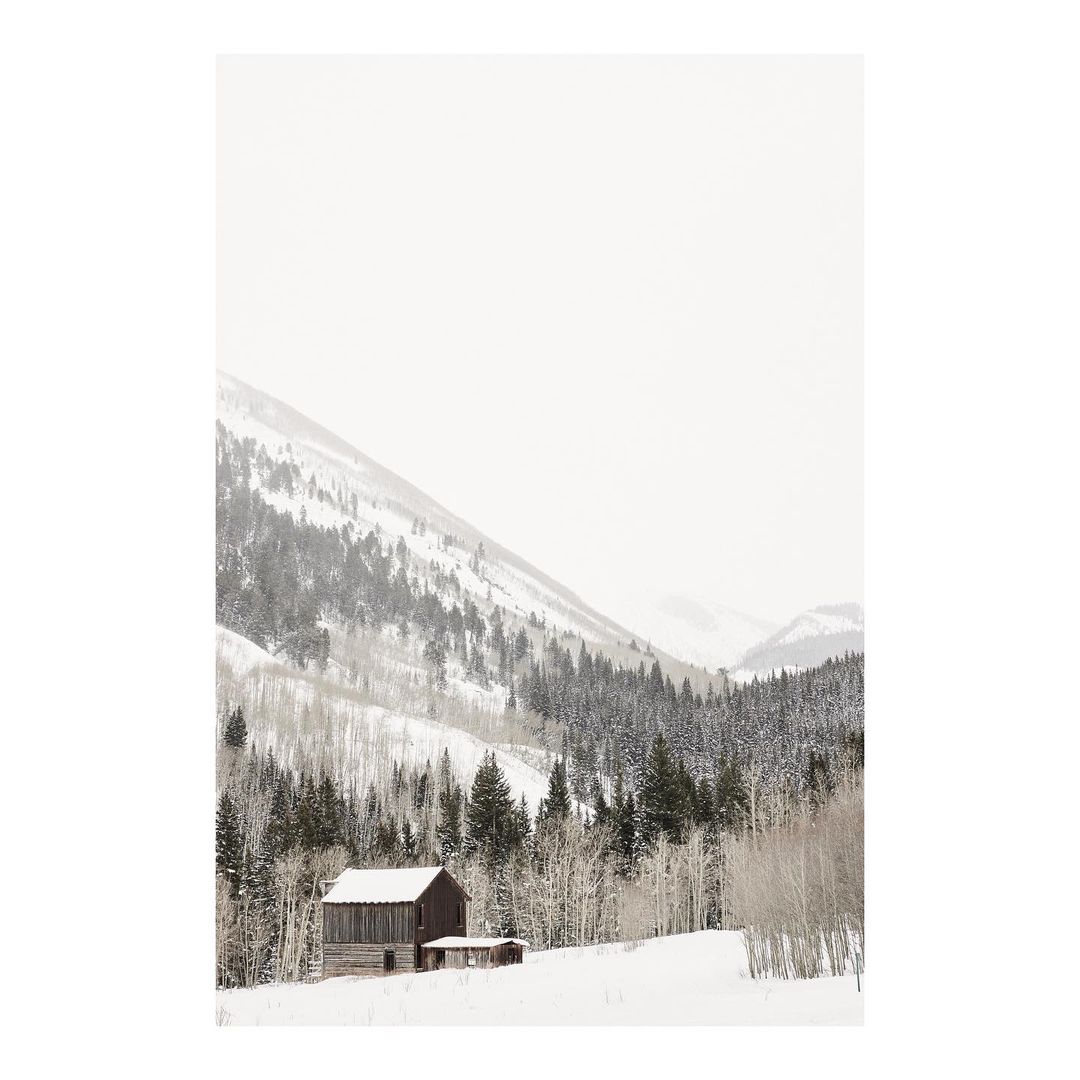Snow-covered barn sits amidst pine trees and snow-dusted mountains, creating a serene winter landscape.