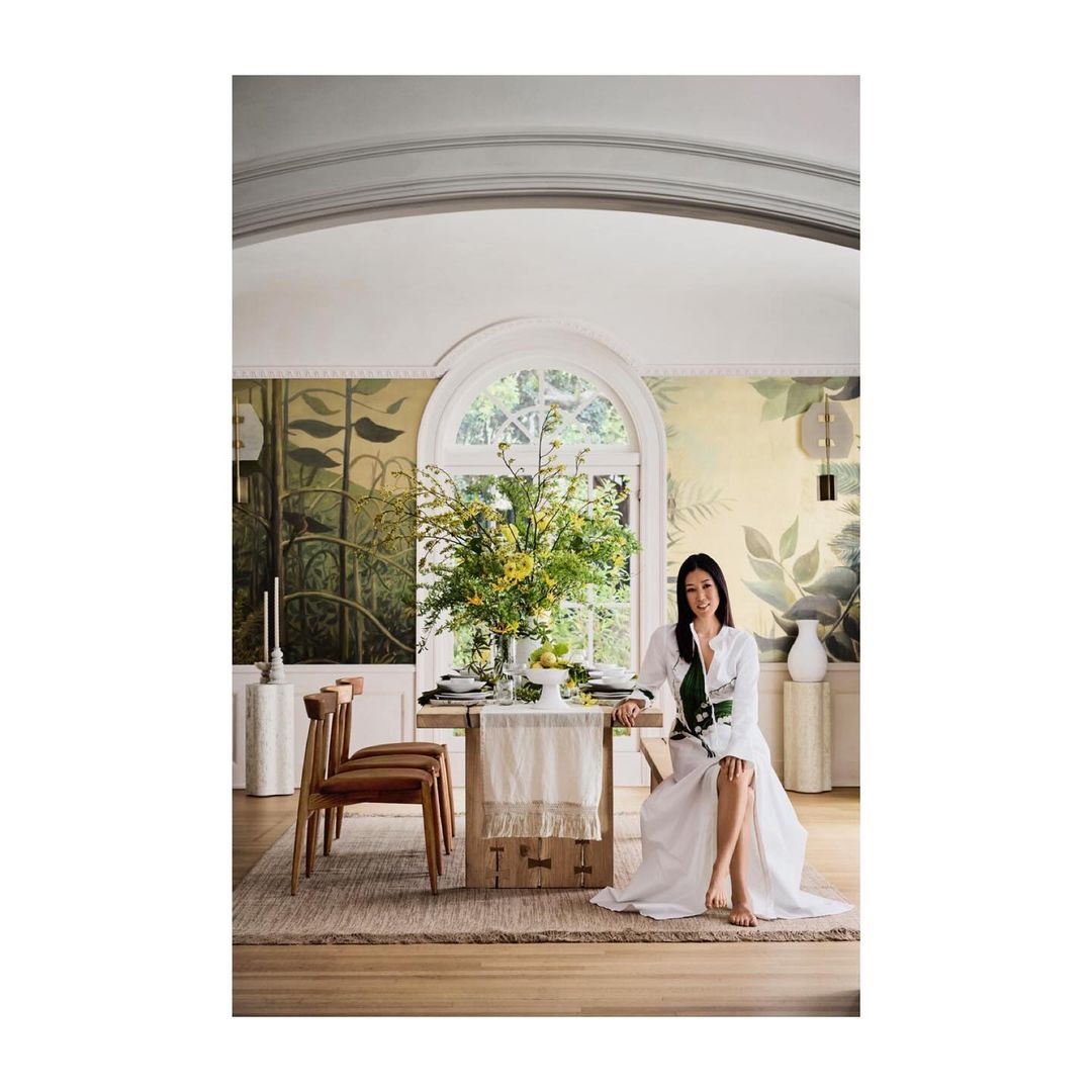 Woman in white dress poses in elegant dining room with floral centerpiece on wooden table, chairs, and botanical wallpaper.