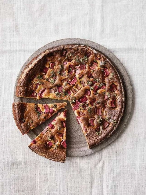 Overhead shot of a rhubarb cake with thyme on a round gray plate with two slices cut out, placed on a white linen tablecloth.