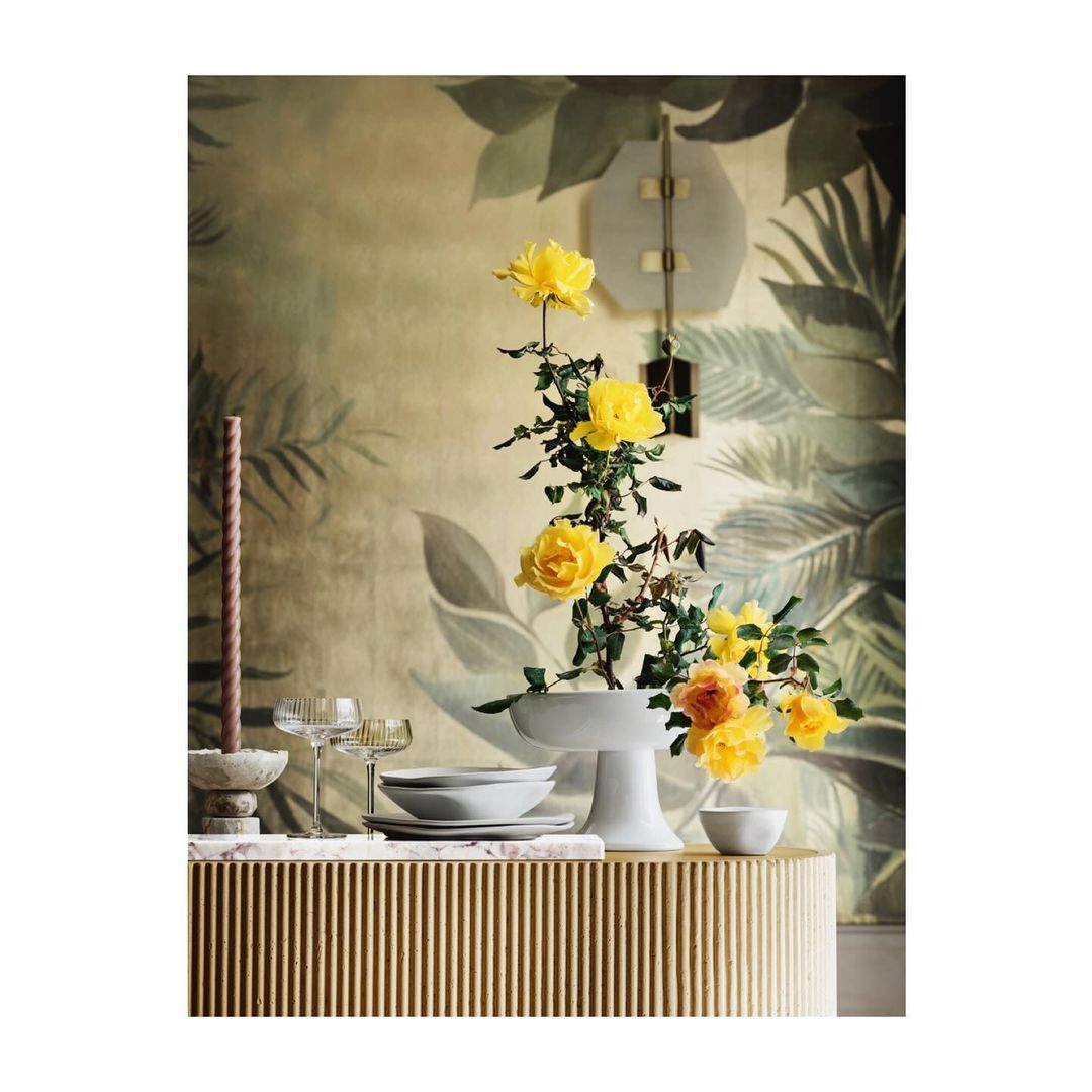Still life of yellow roses in a white vase on a fluted table with champagne glasses and a candle, set against a floral wall.