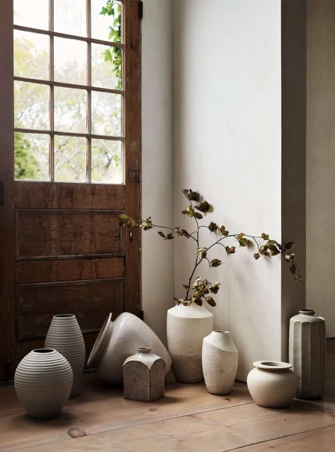 Still life featuring assorted neutral-toned vases on a wooden floor against a rustic door and cream wall, creating a minimalist indoor setting.