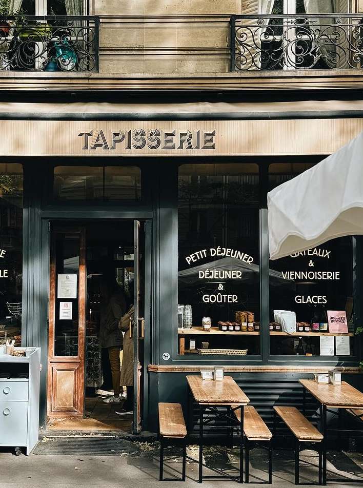 Exterior of a Parisian cafe with a dark-green facade, wooden tables and benches, and a sign reading 'Tapisserie' above the entrance.