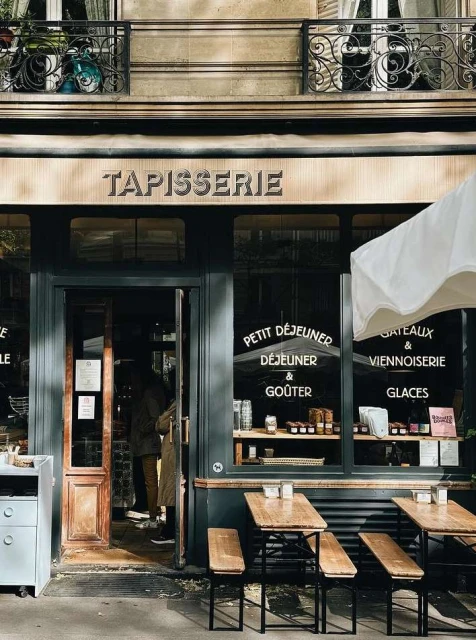 Exterior of a Parisian cafe with a dark-green facade, wooden tables and benches, and a sign reading 'Tapisserie' above the entrance.