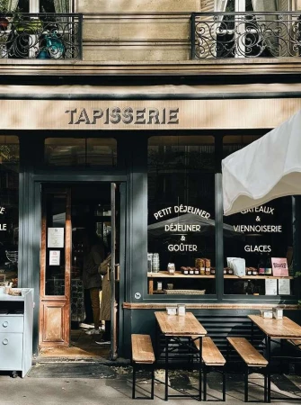 Exterior of a Parisian cafe with a dark-green facade, wooden tables and benches, and a sign reading 'Tapisserie' above the entrance.