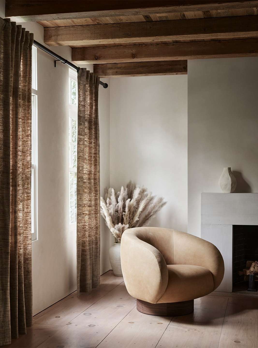 Interior design featuring a neutral-toned lounge chair, pampas grass in a vase, curtains, and a fireplace in a naturally lit room.