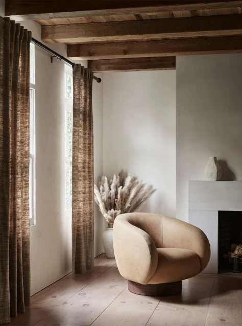 Interior design featuring a neutral-toned lounge chair, pampas grass in a vase, curtains, and a fireplace in a naturally lit room.