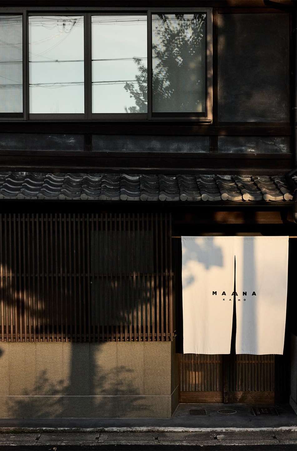Exterior of a Japanese building featuring dark wood, windows, a curtain with text, and tree shadows on a sunny day.