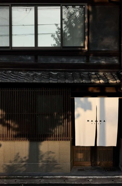 Exterior of a Japanese building featuring dark wood, windows, a curtain with text, and tree shadows on a sunny day.