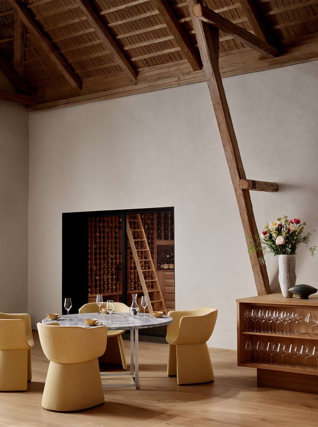 Interior of a dining room featuring a marble table with yellow chairs, wooden floors, and a wine cellar in the background.