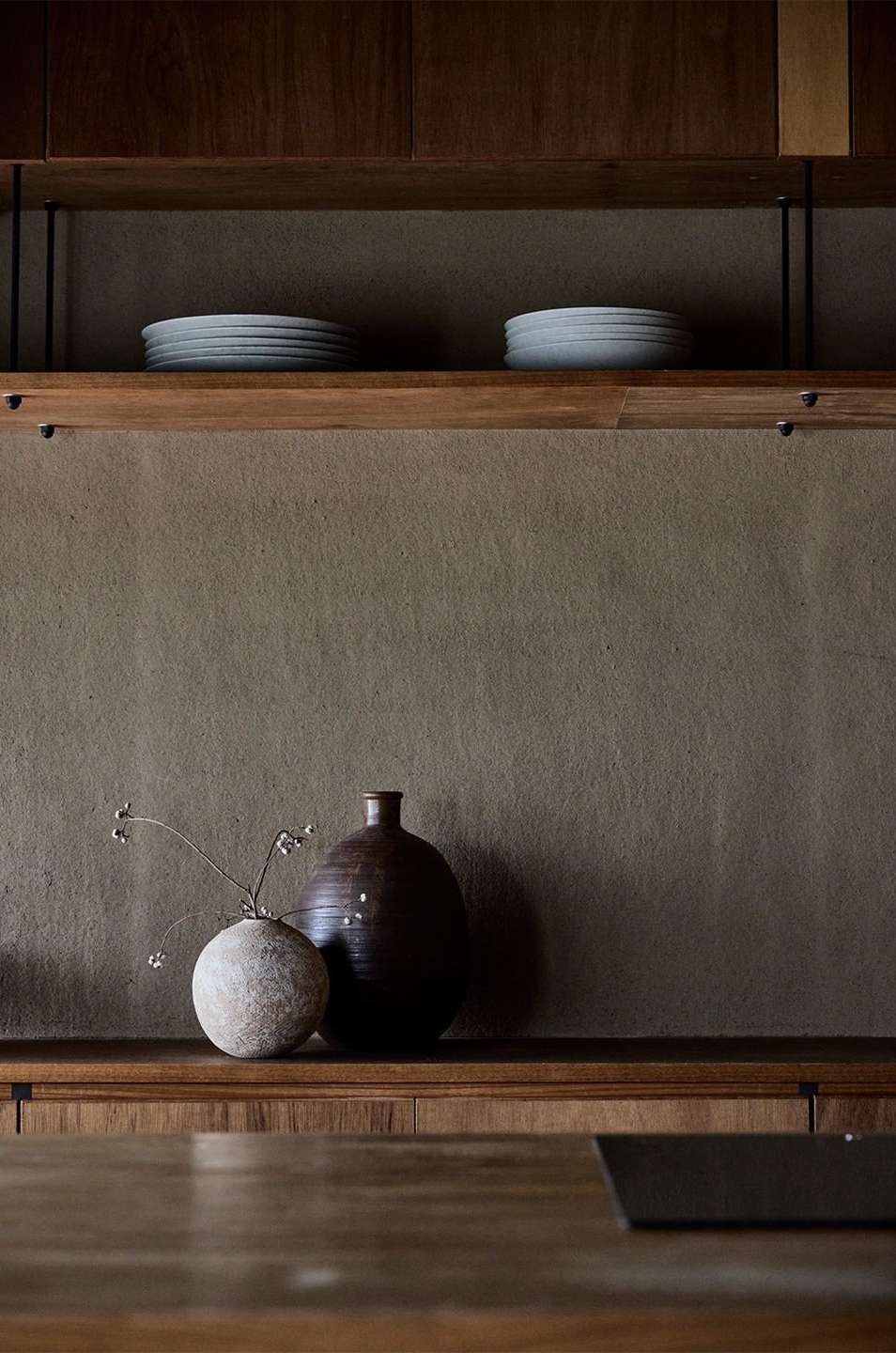 Two vases on a wooden shelf with a stack of white plates, brown wooden cabinets, and a textured grey wall in a neutral interior.