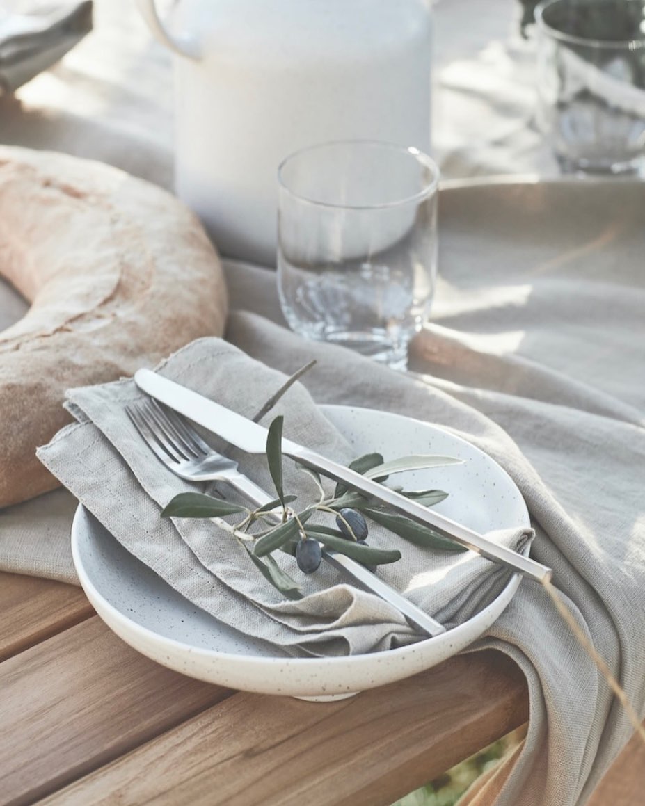 Still life of a table setting with a white plate, silverware, olive branch, glass of water, white pitcher, and bread, in natural light.