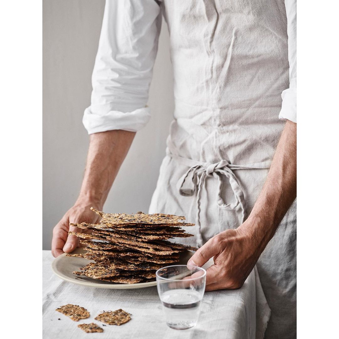 Man in linen apron holding a plate of crispbread next to a glass of water, showcasing a rustic food presentation and healthy eating.