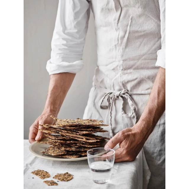 Man in linen apron holding a plate of crispbread next to a glass of water, showcasing a rustic food presentation and healthy eating.