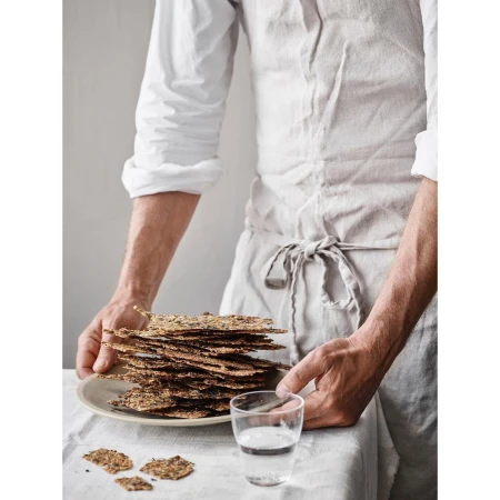 Man in linen apron holding a plate of crispbread next to a glass of water, showcasing a rustic food presentation and healthy eating.