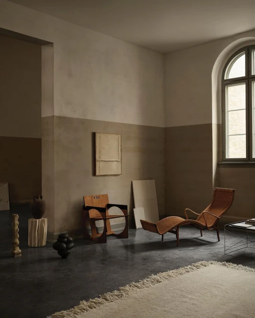 Interior room scene with sculptural wood chairs, brown lounge chair, neutral painting, and textured off-white rug against a gray floor.