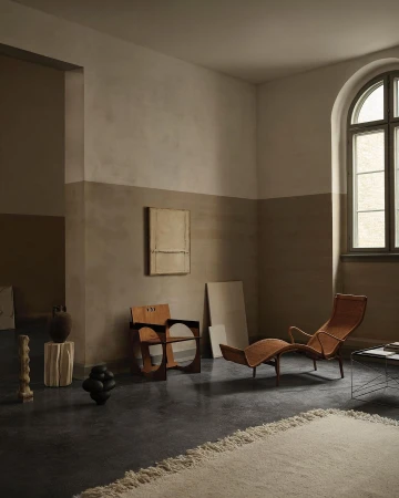 Interior room scene with sculptural wood chairs, brown lounge chair, neutral painting, and textured off-white rug against a gray floor.