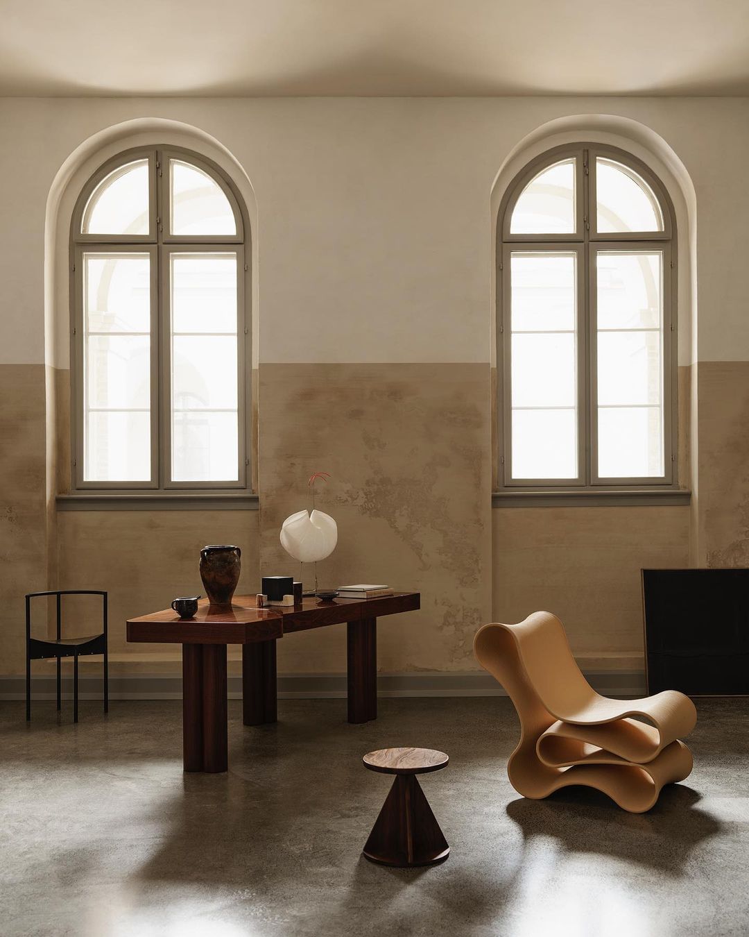 Interior room shot with designer wooden chair, table with objects, chair, conical stool, and arched windows in a neutral color scheme.