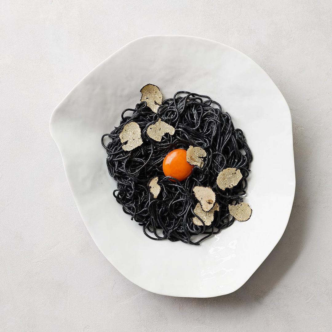 Overhead shot of black spaghetti dish with truffle shavings and an egg yolk in a white asymmetrical bowl on a gray surface.
