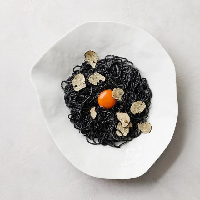Overhead shot of black spaghetti dish with truffle shavings and an egg yolk in a white asymmetrical bowl on a gray surface.