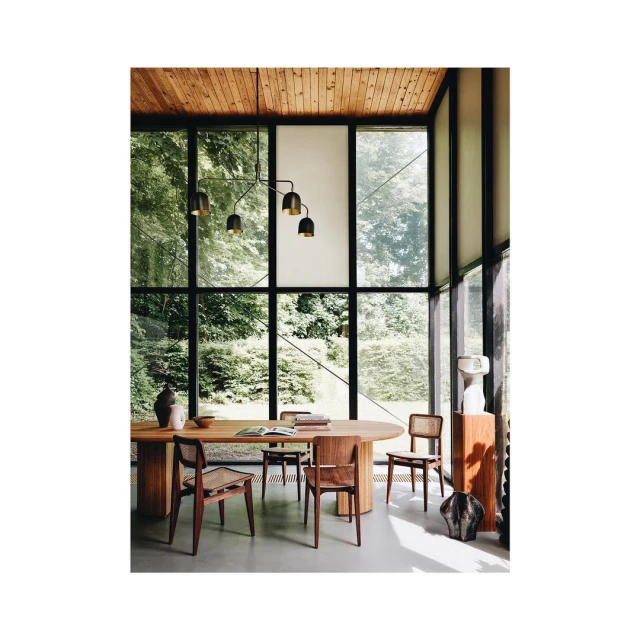 Dining room interior with wood table, chairs, black pendant light and large windows overlooking a green forest exterior setting.