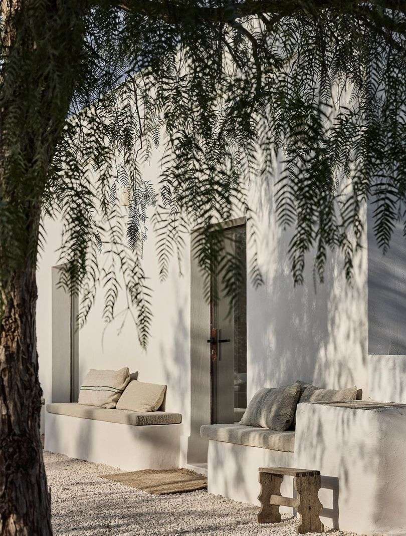 Outdoor seating area with built-in benches, stone floor, and a white building facade, partially shaded by tree branches.