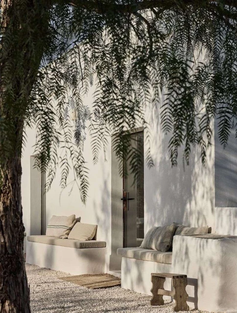 Outdoor seating area with built-in benches, stone floor, and a white building facade, partially shaded by tree branches.