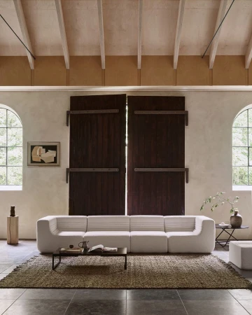 A bright living room interior featuring a modular white sofa, rug, coffee table, and windows providing natural light, creating a minimalist design.