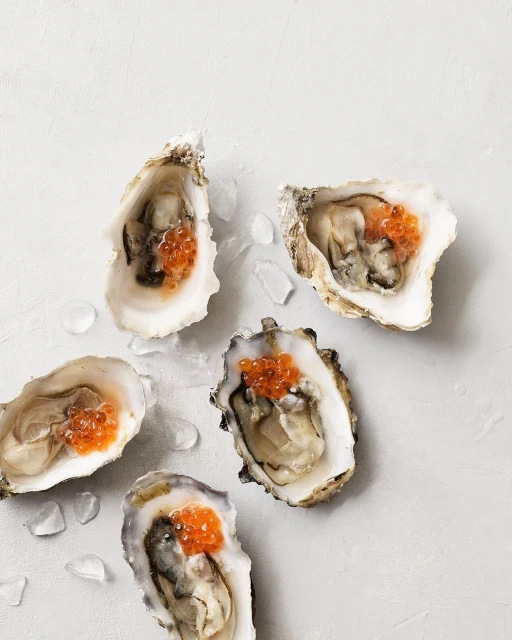 Overhead shot of fresh oysters on the half shell, adorned with red caviar, served on a light grey surface with ice crystals.