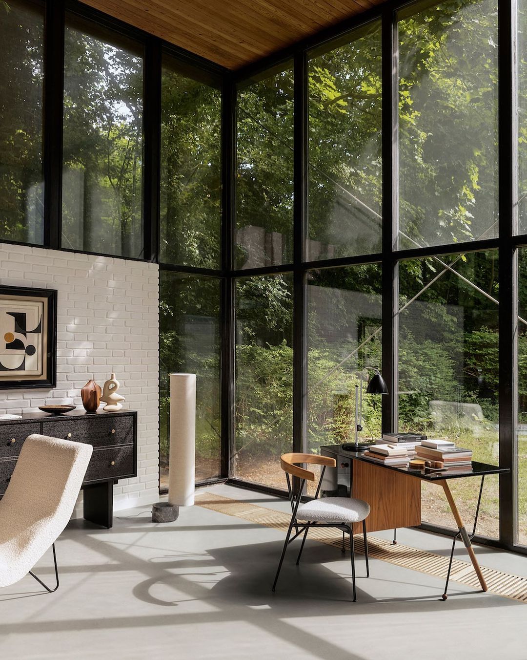 Modern office interior with floor-to-ceiling windows overlooking green trees, featuring a desk, chair, minimalist décor, and natural light.