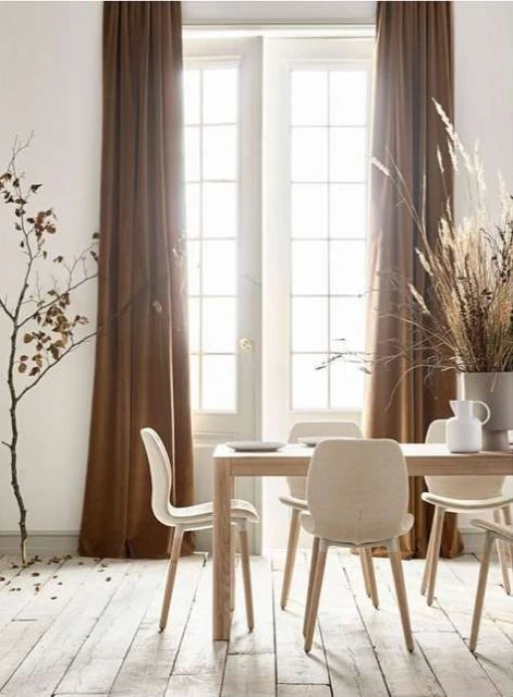 Interior of a dining room with a light wood table and chairs, set against white walls, brown curtains, and a bright window.