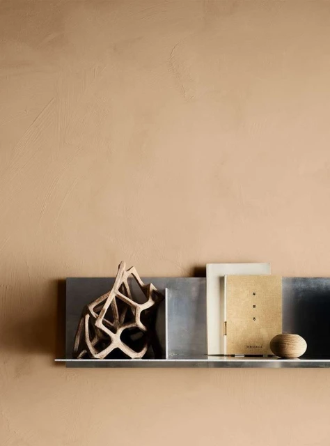 Still life featuring a modern geometric sculpture, books, and a small wooden ball on a metallic shelf against a textured beige wall.