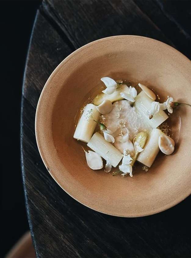 Overhead shot of an artistic food presentation: peeled white asparagus, edible flowers, and a light sauce in a shallow ceramic bowl.