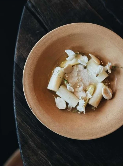 Overhead shot of an artistic food presentation: peeled white asparagus, edible flowers, and a light sauce in a shallow ceramic bowl.