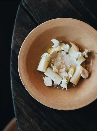 Overhead shot of an artistic food presentation: peeled white asparagus, edible flowers, and a light sauce in a shallow ceramic bowl.