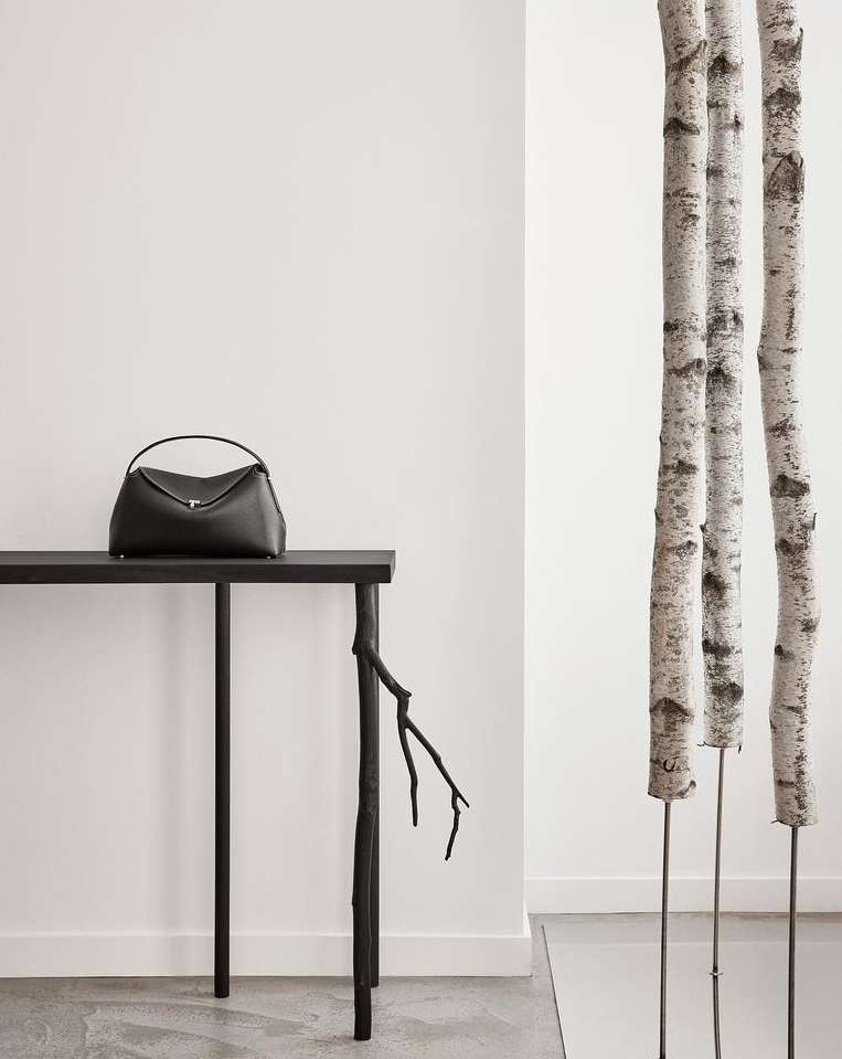 Elegant black leather handbag displayed on a sleek black table with decorative branch leg, complemented by birch tree trunks against a white wall.