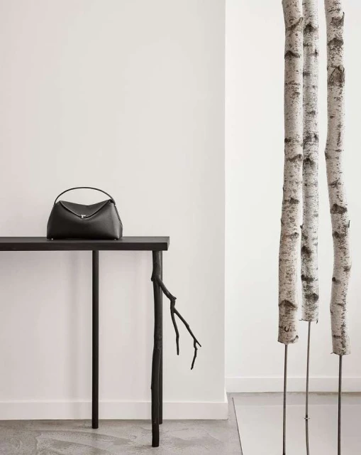 Elegant black leather handbag displayed on a sleek black table with decorative branch leg, complemented by birch tree trunks against a white wall.