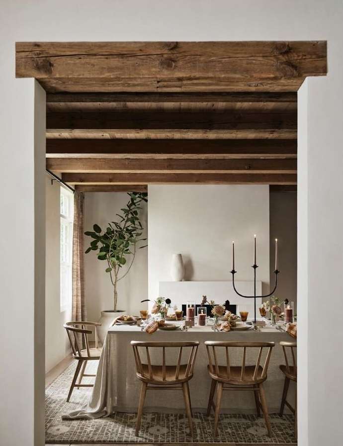 Dining room interior with a table set for a meal, wooden chairs, neutral color scheme, and natural light streaming through a window.