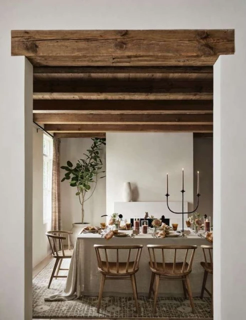 Dining room interior with a table set for a meal, wooden chairs, neutral color scheme, and natural light streaming through a window.