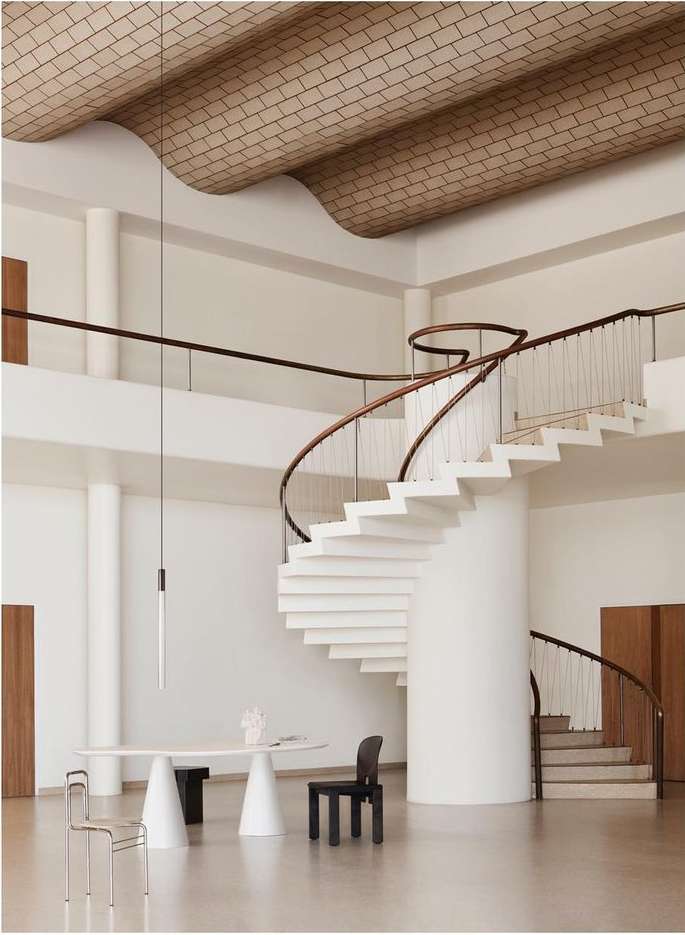 Bright, airy interior with a white spiral staircase, sculptural table and chairs, modern pendant light, and geometric textured ceiling.