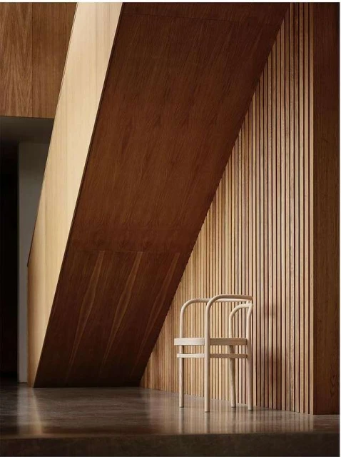 Minimalist interior with a light wood chair against a slatted wood wall, accented by strong diagonal lines from overhead architectural elements.
