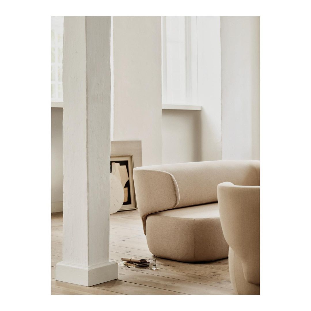Living room interior featuring a beige curved sofa and armchair, complemented by an abstract framed print and a white architectural pillar.
