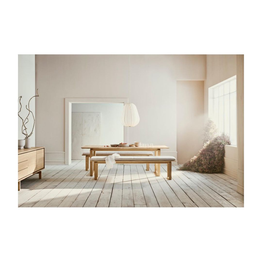 Neutral toned dining space featuring a light wood table, benches, pendant lamp, and modern sideboard in a sunlit room. Interior design.