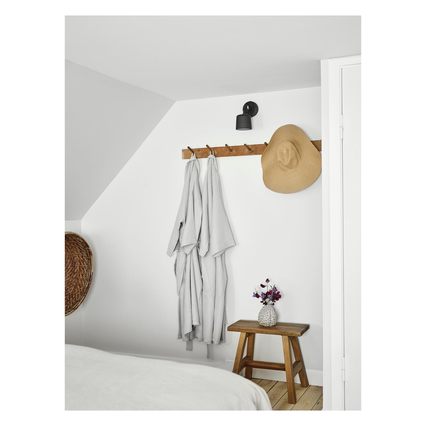 Bedroom interior with a bed, wooden stool topped with a vase of flowers, a light fixture, and items hanging on wall hooks.