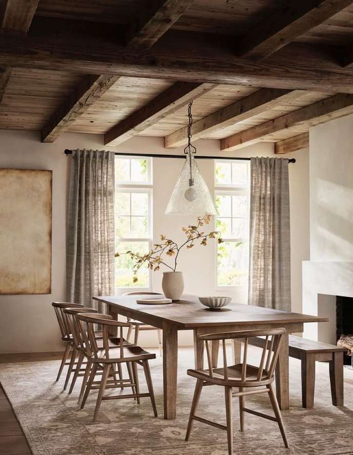 Interior of a dining room with a wooden table and chairs, a decorative vase, wood ceiling beams, and natural light from windows.