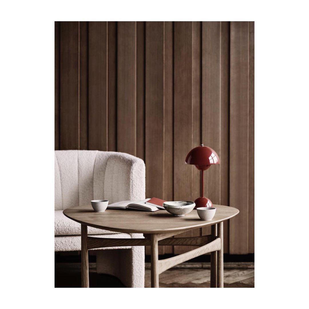 Minimalist interior featuring a red table lamp on a round wooden table, next to a white couch and a wood-panelled wall.
